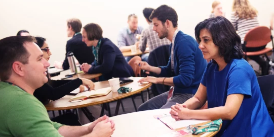 A room full of people meeting one on one. The focus of the image is a woman in blue dress at a table meeting with a man in a green shirt.