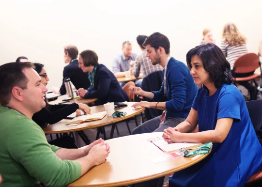 6015acbbd11b9706af971456_Image-2-e1561487612267 A room full of people meeting one on one. The focus of the image is a woman in blue dress at a table meeting with a man in a green shirt.