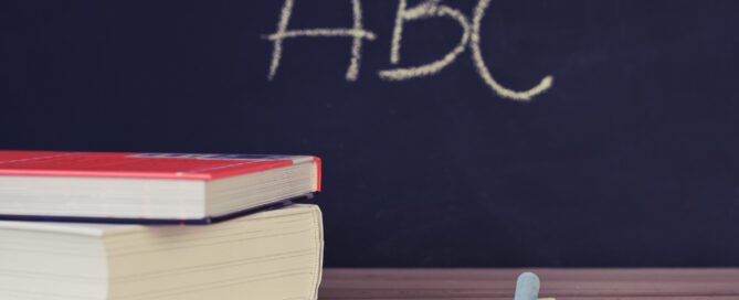 TwoBooks Beside Two Chalks on a desk in front of a blackboard. Om the blackboard are the letters